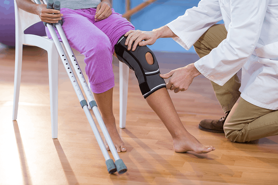 Woman with crutches and knee support wrap being examined by medical professional