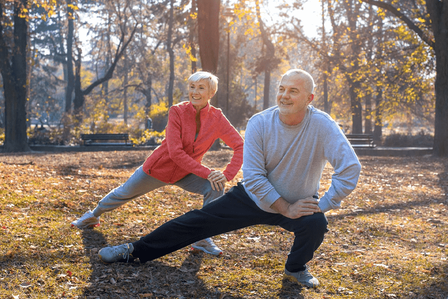 senior couple in wooded park doing standing knee stretches